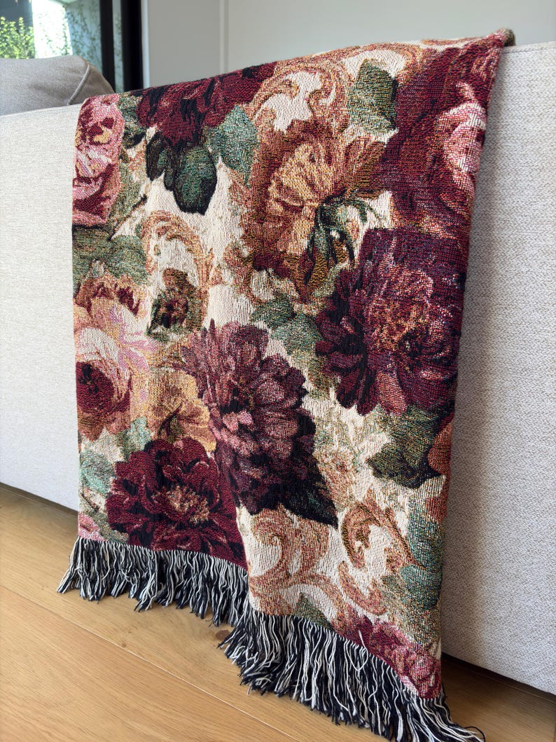 May include: A floral tapestry throw blanket with a cream background and a pattern of burgundy, pink, and green flowers. The blanket has a fringe trim with black and white threads. The blanket is draped over a light gray sofa.