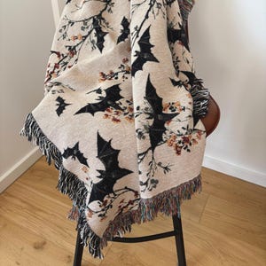 May include: Beige woven throw blanket featuring a repeating pattern of black bats and floral branches. The blanket has a fringed edge with a mix of colors. The throw is draped over a chair with a black metal frame.
