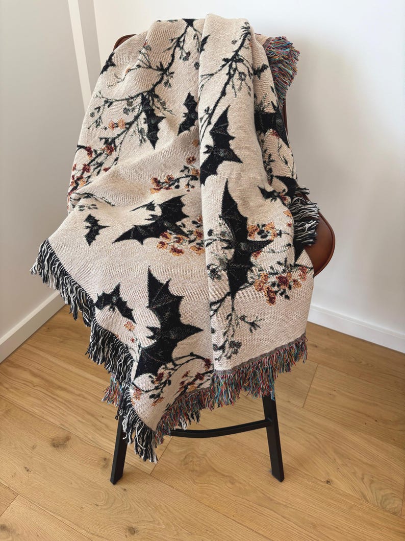 May include: Beige woven throw blanket featuring a repeating pattern of black bats and floral branches. The blanket has a fringed edge with a mix of colors. The throw is draped over a chair with a black metal frame.