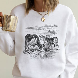 K&ouml;nnte beinhalten: Wei&szlig;es Sweatshirt mit einer Schwarzwei&szlig;-Illustration von vier K&uuml;hen auf einer Weide. Die K&uuml;he trinken aus einem Bach. Die Illustration ist im Vintage-Stil gehalten.