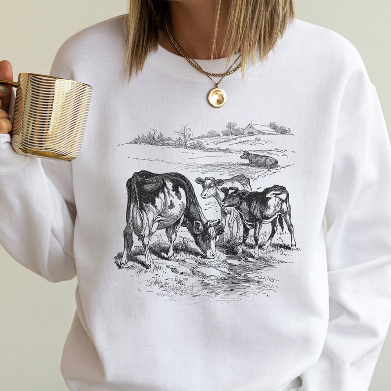 K&ouml;nnte beinhalten: Wei&szlig;es Sweatshirt mit einer Schwarzwei&szlig;-Illustration von vier K&uuml;hen auf einer Weide. Die K&uuml;he trinken aus einem Bach. Die Illustration ist im Vintage-Stil gehalten.