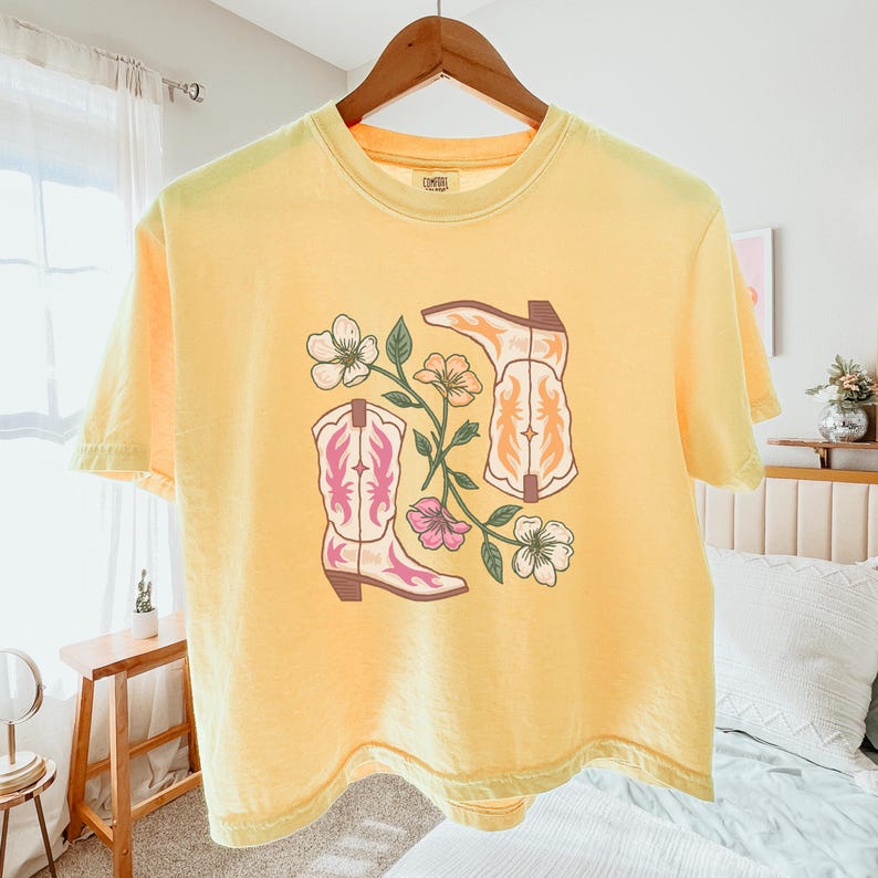 May include: A yellow short-sleeved t-shirt with a floral design and two pairs of cowboy boots. The design features pink, white, and yellow flowers and green leaves. The boots are white with brown accents. The t-shirt has a distressed look and is made of a soft, comfortable fabric.