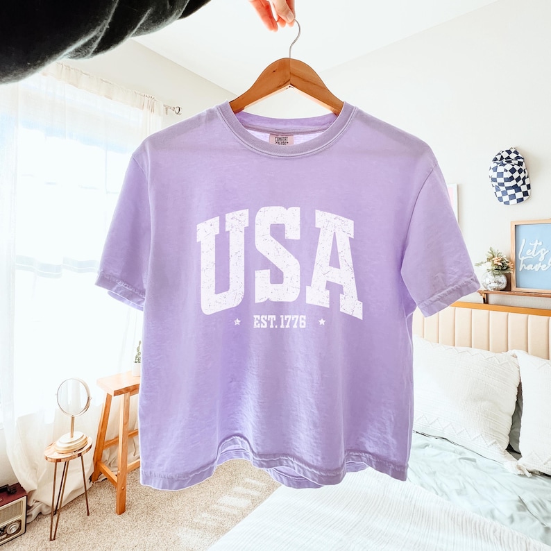 Pu&ograve; includere: Una maglietta lilla chiaro con la scritta "USA" a blocchi bianchi. Sotto la parola "USA" c'&egrave; la scritta "EST. 1776" con una stella su ogni lato. La maglietta &egrave; appesa a una gruccia di legno.