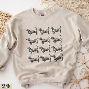 Peut inclure: Un sweat-shirt beige avec un motif r&eacute;p&eacute;t&eacute; de chiens teckel noirs. Le sweat-shirt a un col rond et des manches longues.