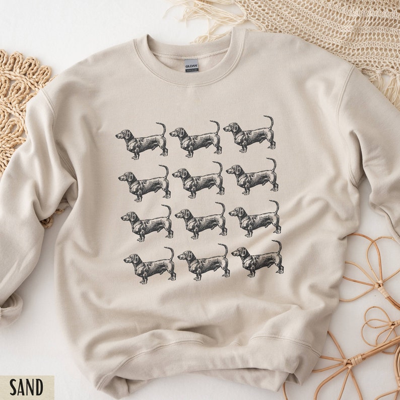 Peut inclure: Un sweat-shirt beige avec un motif r&eacute;p&eacute;t&eacute; de chiens teckel noirs. Le sweat-shirt a un col rond et des manches longues.