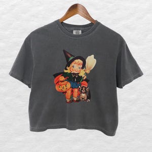 May include: Dark gray t-shirt featuring a vintage-style illustration of a young witch holding a pumpkin and broom, with a black cat. The witch is wearing a black hat and cape. The shirt is hanging on a wooden hanger.