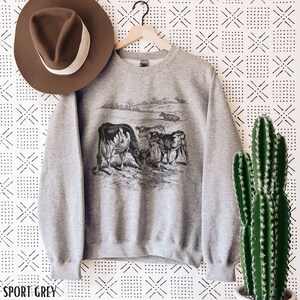 K&ouml;nnte beinhalten: Ein graues Sweatshirt mit einer Vintage-Illustration von K&uuml;hen auf einer Weide. Das Sweatshirt hat den Schriftzug "SPORT GREY" unten aufgedruckt.