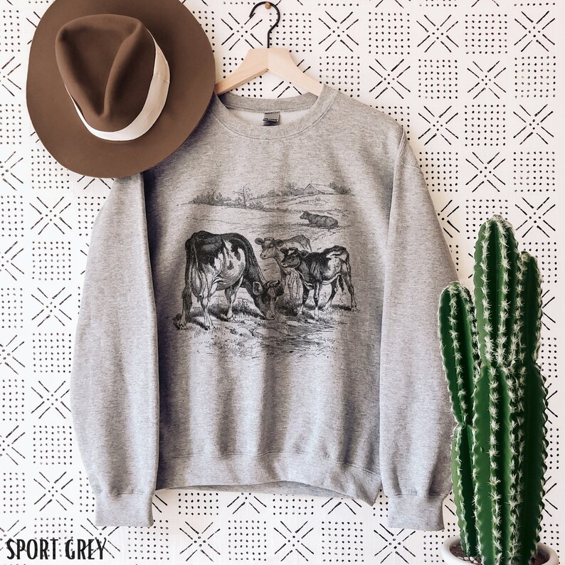 K&ouml;nnte beinhalten: Ein graues Sweatshirt mit einer Vintage-Illustration von K&uuml;hen auf einer Weide. Das Sweatshirt hat den Schriftzug "SPORT GREY" unten aufgedruckt.