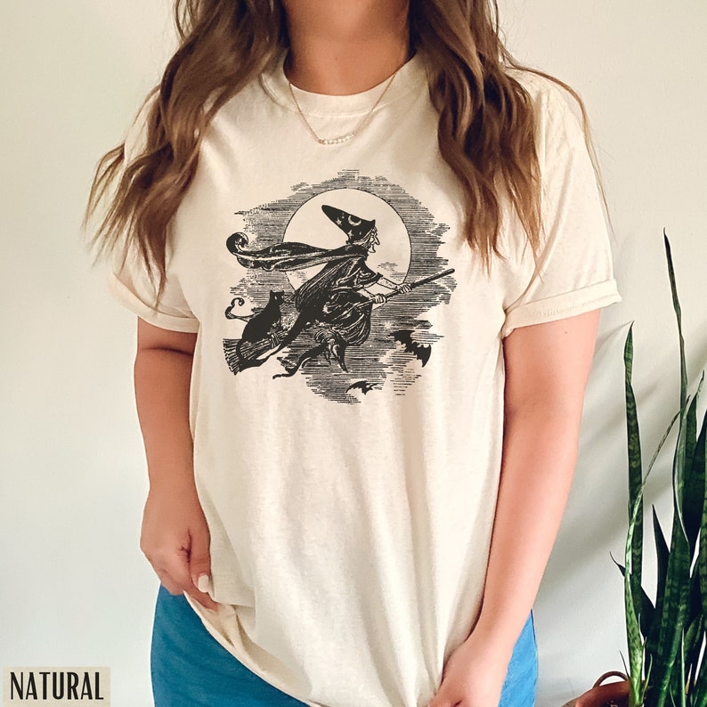 May include: Cream-colored t-shirt featuring a black and white illustration of a witch on a broomstick, flying in front of a full moon, with a black cat and bats. The word "NATURAL" is printed at the bottom left.