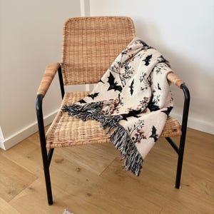 May include: A woven throw blanket with a Halloween theme draped over a wicker chair. The blanket features a cream background with black bats and floral accents. The blanket has a black fringe.