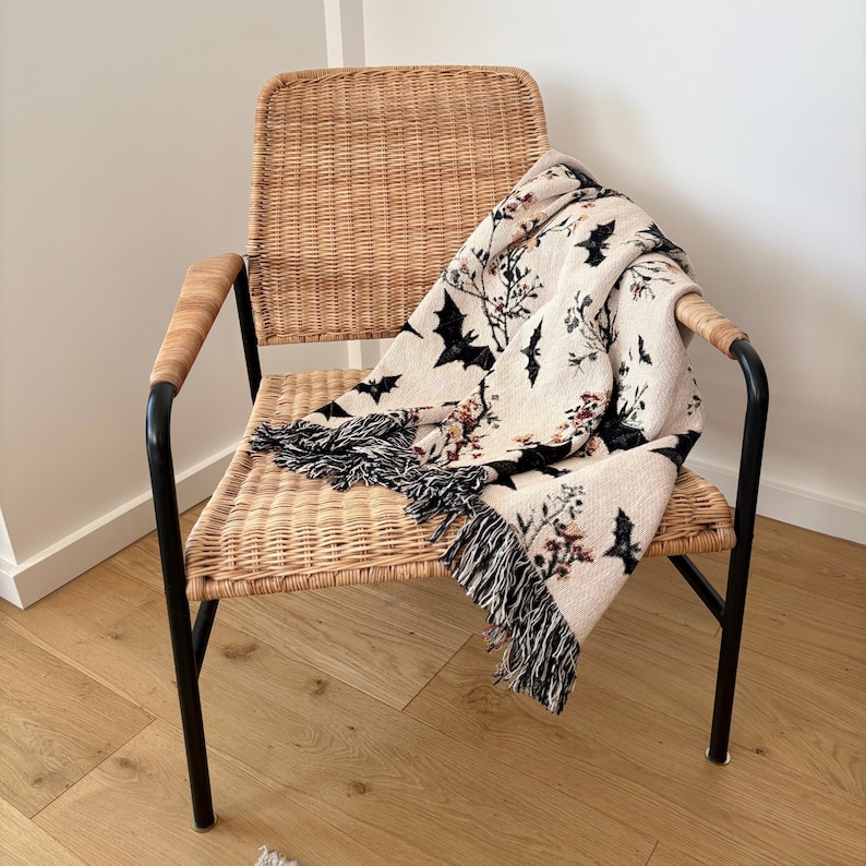May include: A woven throw blanket with a Halloween theme draped over a wicker chair. The blanket features a cream background with black bats and floral accents. The blanket has a black fringe.