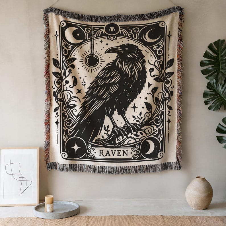 May include: A beige throw blanket with a black raven illustration. The raven is perched on a branch within a decorative frame, with the word "RAVEN" at the bottom. The blanket has a fringed edge and is hanging on a wall.