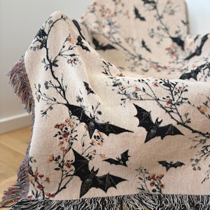 May include: A beige throw blanket with a repeating pattern of black bats and floral branches. The blanket has a fringed edge with a mix of colors. The blanket is draped over a chair.