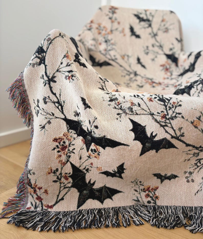 May include: A beige throw blanket with a repeating pattern of black bats and floral branches. The blanket has a fringed edge with a mix of colors. The blanket is draped over a chair.