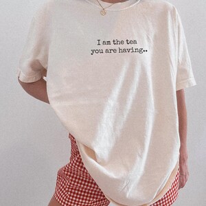 May include: Cream-colored t-shirt with the text "I am the tea you are having.." in black. The shirt is paired with red and white checkered shorts. The outfit is casual and comfortable.