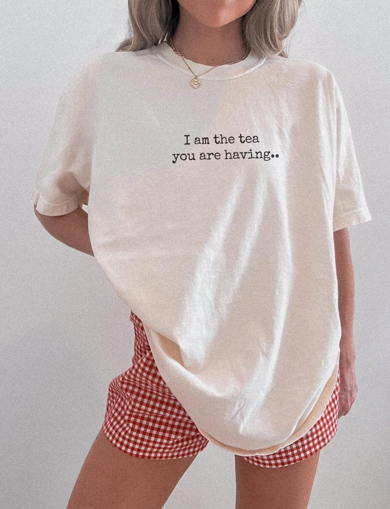 May include: Cream-colored t-shirt with the text "I am the tea you are having.." in black. The shirt is paired with red and white checkered shorts. The outfit is casual and comfortable.