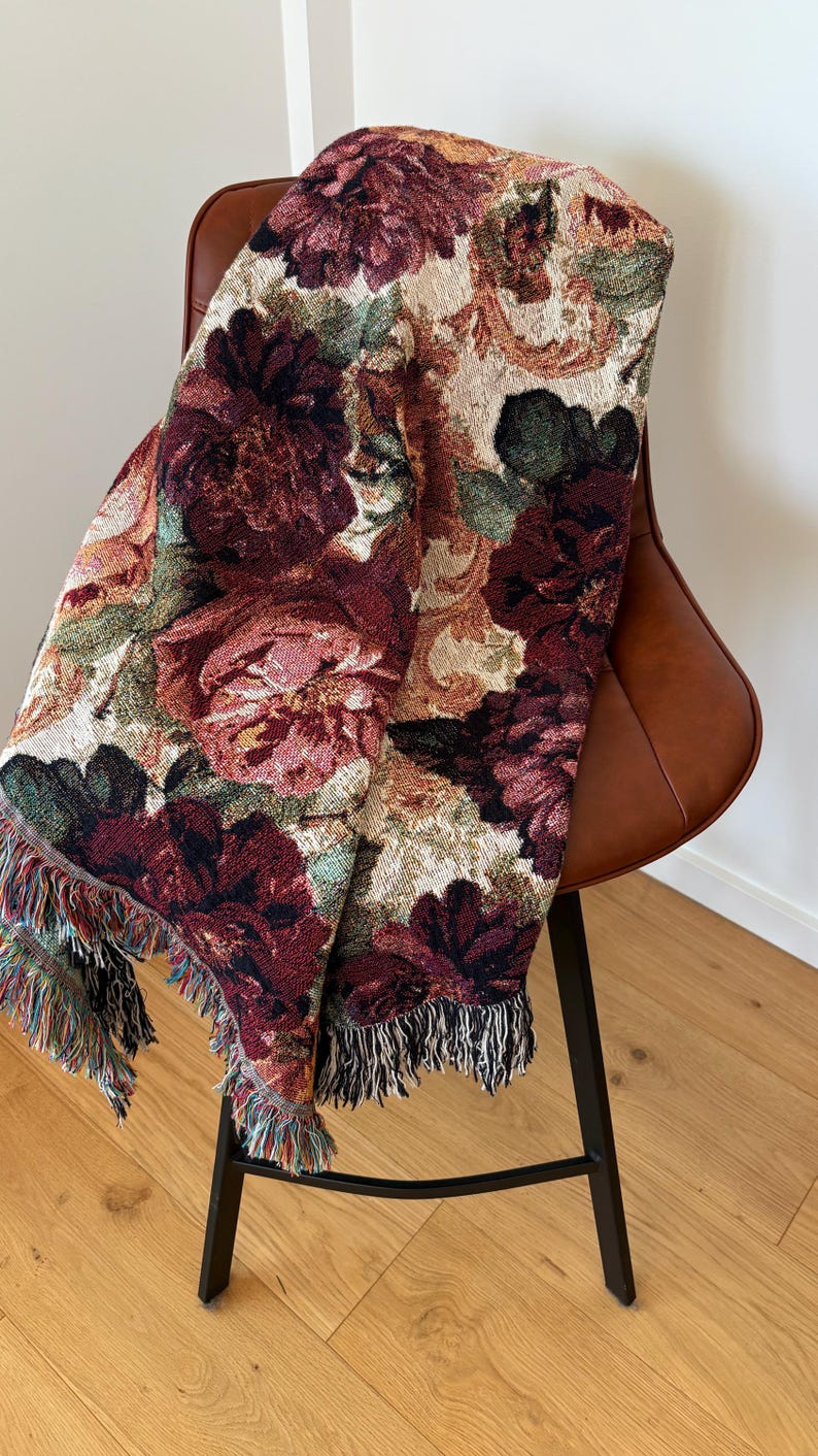 May include: A floral tapestry throw blanket draped over a brown leather chair. The blanket features a pattern of large burgundy, pink, and cream-colored flowers with green leaves. The blanket has a fringed edge with multiple colors.