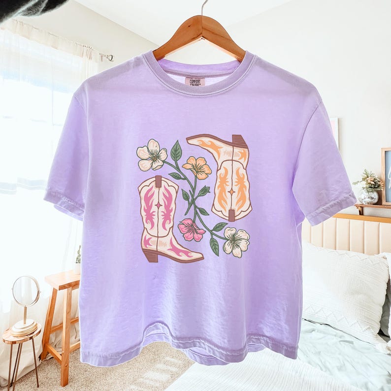May include: A lilac t-shirt with a graphic of two cowboy boots and floral accents in pink, orange, and white. The boots have decorative stitching. The shirt is displayed on a wooden hanger.