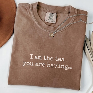 May include: A brown Comfort Colors t-shirt with the text "I am the tea you are having." A silver necklace with a small pendant is draped across the shirt. The shirt is on a white surface.