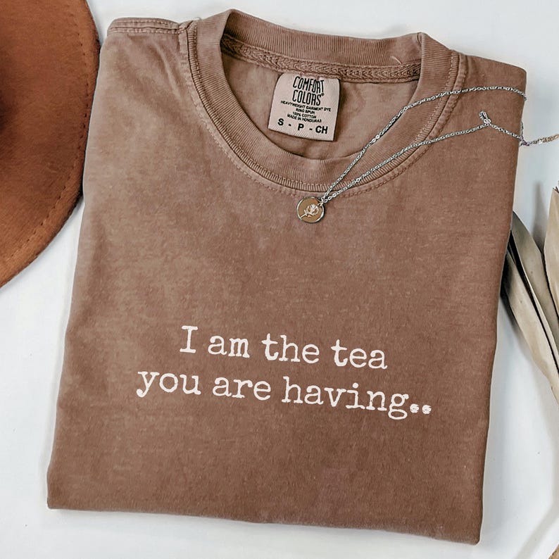 May include: A brown Comfort Colors t-shirt with the text "I am the tea you are having." A silver necklace with a small pendant is draped across the shirt. The shirt is on a white surface.