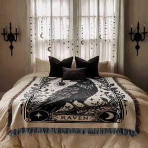 May include: A cream-colored throw blanket with a black raven illustration and the word "RAVEN". The blanket has a fringed edge and is displayed on a bed with black pillows and decorative wall sconces.