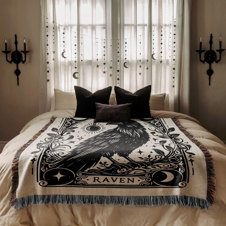 May include: A cream-colored throw blanket with a black raven illustration and the word "RAVEN". The blanket has a fringed edge and is displayed on a bed with black pillows and decorative wall sconces.