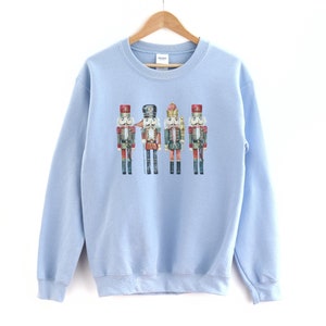 May include: A light blue sweatshirt featuring a watercolor illustration of four toy nutcracker soldiers. The nutcrackers are dressed in red, black, green, and gold uniforms with detailed facial features and hats. The sweatshirt is hanging on a wooden hanger.
