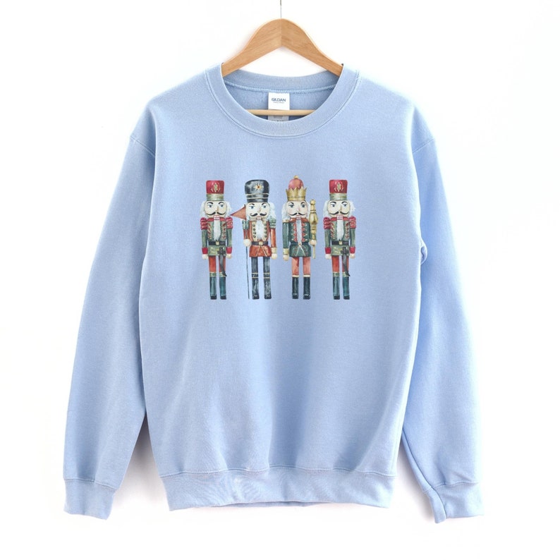 May include: A light blue sweatshirt featuring a watercolor illustration of four toy nutcracker soldiers. The nutcrackers are dressed in red, black, green, and gold uniforms with detailed facial features and hats. The sweatshirt is hanging on a wooden hanger.