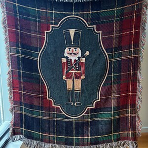 May include: A woven throw blanket with a plaid pattern in shades of red, green, and navy blue. The center features a framed image of a nutcracker soldier in red and gold. The blanket has a fringed edge.