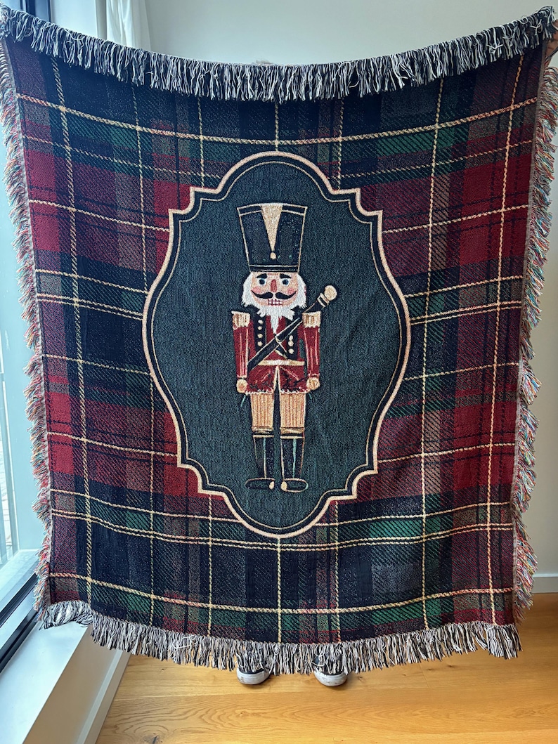 May include: A woven throw blanket with a plaid pattern in shades of red, green, and navy blue. The center features a framed image of a nutcracker soldier in red and gold. The blanket has a fringed edge.