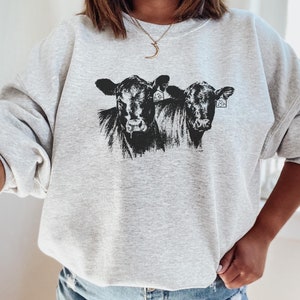 K&ouml;nnte beinhalten: Ein hellgraues Sweatshirt mit einer schwarz-wei&szlig;en Grafik von zwei K&uuml;hen. Die K&uuml;he haben Ohrmarken. Das Sweatshirt hat lange &Auml;rmel und einen Rundhalsausschnitt.