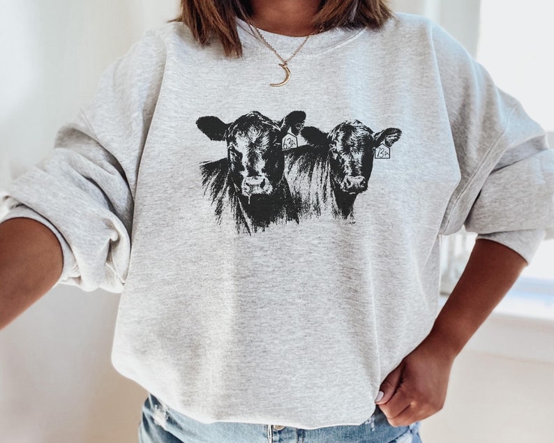 K&ouml;nnte beinhalten: Ein hellgraues Sweatshirt mit einer schwarz-wei&szlig;en Grafik von zwei K&uuml;hen. Die K&uuml;he haben Ohrmarken. Das Sweatshirt hat lange &Auml;rmel und einen Rundhalsausschnitt.