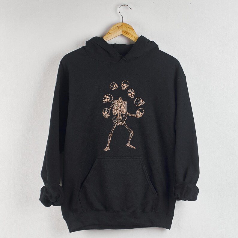 May include: Black hooded sweatshirt featuring a graphic print of a skeleton juggling skulls. The skeleton is standing with its arms outstretched, holding skulls in each hand. The skulls are arranged in a circle around the skeleton's head.