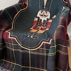 May include: A woven throw blanket featuring a plaid pattern in black, red, and green. The center showcases a detailed image of a nutcracker soldier in red and gold, set against a dark gray background. The blanket has a fringed edge.