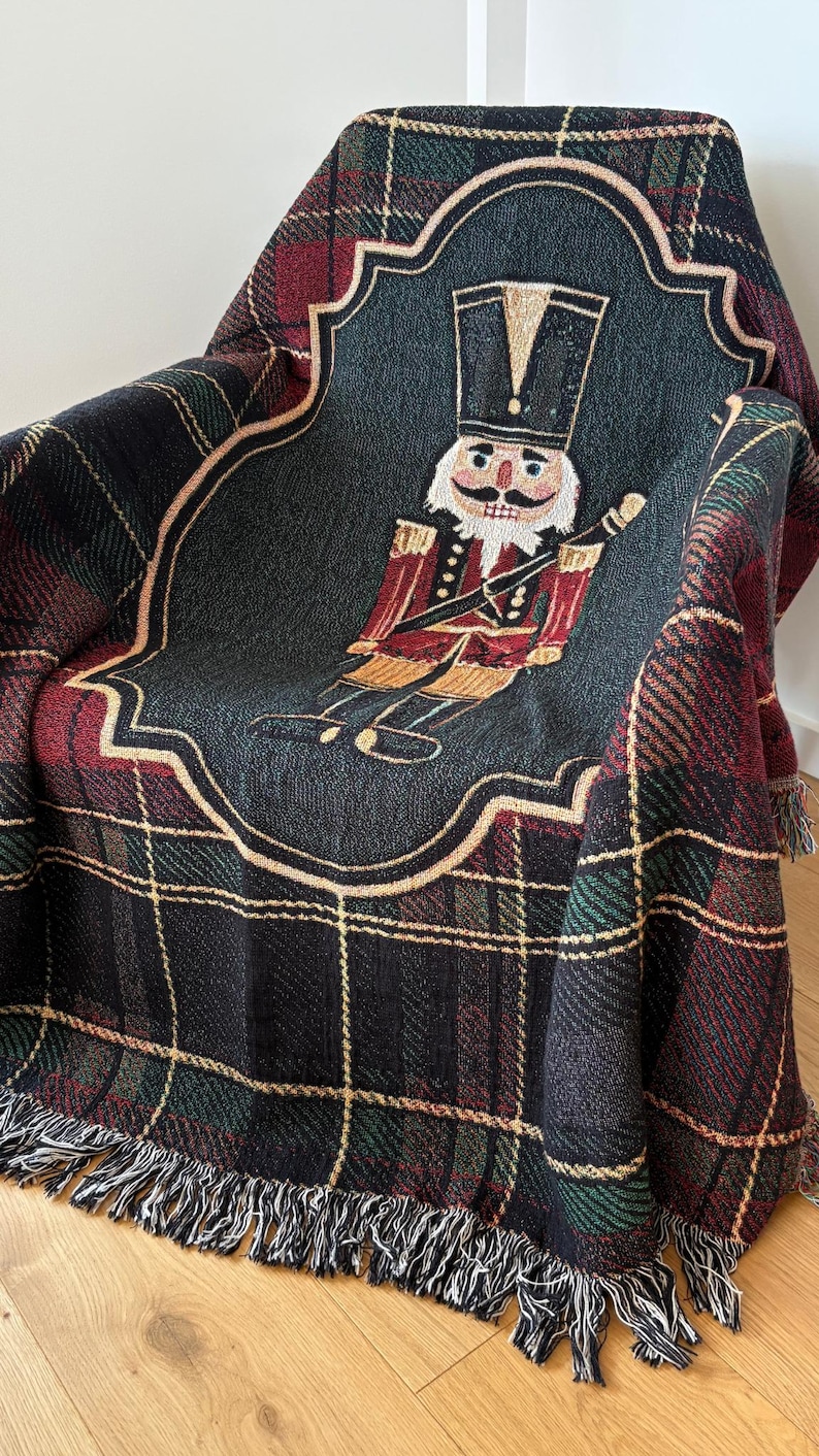 May include: A woven throw blanket featuring a plaid pattern in black, red, and green. The center showcases a detailed image of a nutcracker soldier in red and gold, set against a dark gray background. The blanket has a fringed edge.
