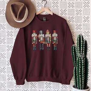 May include: Burgundy crewneck sweatshirt featuring a watercolor illustration of four nutcrackers. The nutcrackers are dressed in red, green, and black uniforms with gold accents. A brown hat hangs on a wooden hanger above the sweatshirt.
