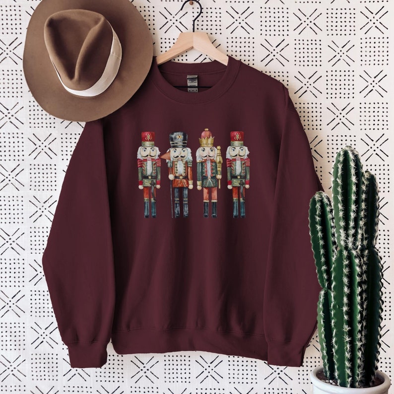 May include: Burgundy crewneck sweatshirt featuring a watercolor illustration of four nutcrackers. The nutcrackers are dressed in red, green, and black uniforms with gold accents. A brown hat hangs on a wooden hanger above the sweatshirt.