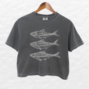 Puede incluir: Una camiseta gris oscuro recortada con un gr&aacute;fico de tres peces estilizados impresos en la parte delantera.