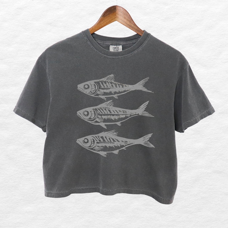 Puede incluir: Una camiseta gris oscuro recortada con un gr&aacute;fico de tres peces estilizados impresos en la parte delantera.