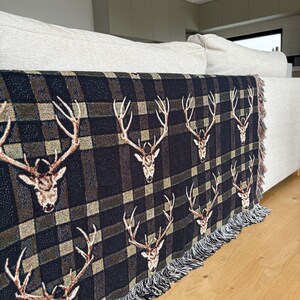 K&ouml;nnte beinhalten: Eine schwarz-braune karierte Decke mit einem sich wiederholenden Muster von Hirschk&ouml;pfen. Die Decke mit Fransen ist &uuml;ber ein helles Sofa drapiert. Die Farbpalette umfasst Braun-, Schwarz- und Beiget&ouml;ne. Geeignet f&uuml;r ein Sofa.