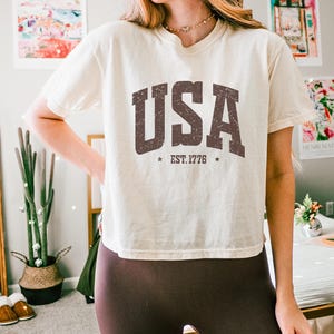 Pu&ograve; includere: T-shirt color crema con la scritta "USA" in lettere marroni consumate. Sotto, "EST. 1776" con stelle su entrambi i lati. La maglietta &egrave; abbinata a leggings marroni scuri.
