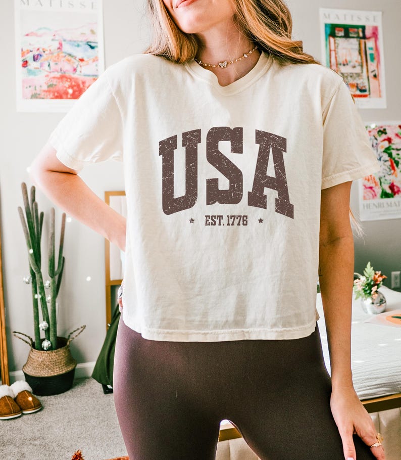 Pu&ograve; includere: T-shirt color crema con la scritta "USA" in lettere marroni consumate. Sotto, "EST. 1776" con stelle su entrambi i lati. La maglietta &egrave; abbinata a leggings marroni scuri.