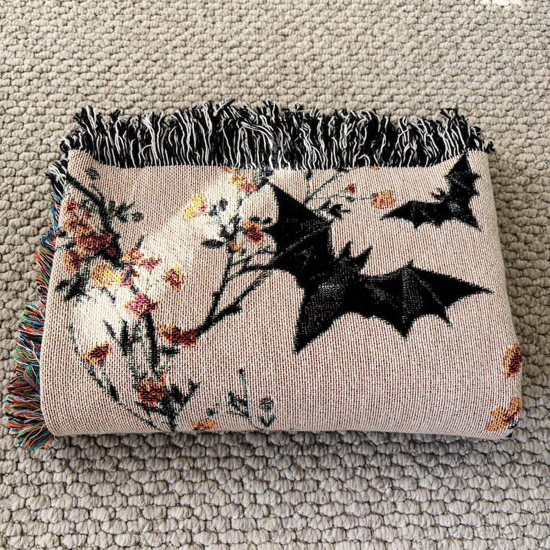 May include: Beige woven throw blanket featuring black bats and floral branches. The blanket has a fringed edge in black and a multicolored fringe on one end. The design includes orange, yellow, and red flowers.