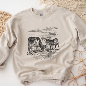 K&ouml;nnte beinhalten: Ein beiger Pullover mit einer Vintage-Illustration von K&uuml;hen auf einer Weide. Die Illustration ist in Schwarzwei&szlig; gehalten und zeigt drei K&uuml;he, darunter ein Kalb.