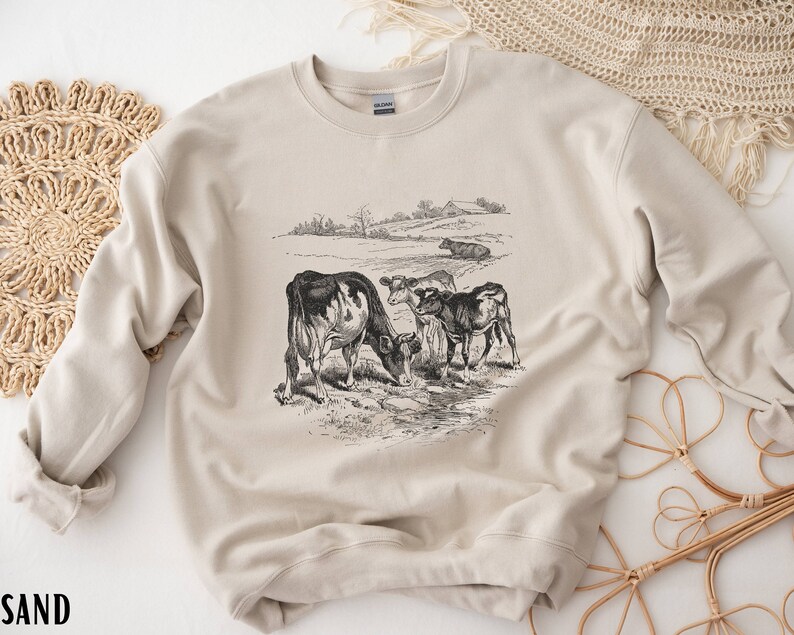 K&ouml;nnte beinhalten: Ein beiger Pullover mit einer Vintage-Illustration von K&uuml;hen auf einer Weide. Die Illustration ist in Schwarzwei&szlig; gehalten und zeigt drei K&uuml;he, darunter ein Kalb.