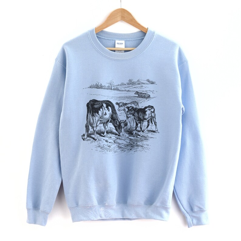 K&ouml;nnte beinhalten: Ein hellblauer Pullover mit einer Schwarzwei&szlig;illustration von K&uuml;hen auf einer Weide. Der Pullover hat einen Rundhalsausschnitt und lange &Auml;rmel.