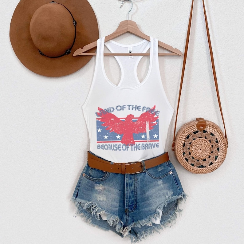 May include: White tank top with a red, white, and blue eagle graphic and the text "Land of the Free Because of the Brave". The tank top is paired with denim shorts and a brown belt.
