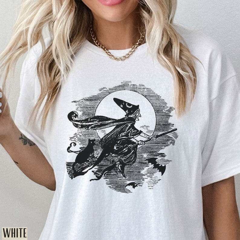May include: White t-shirt featuring a black and white illustration of a witch flying on a broomstick in front of a full moon, with a black cat and bats. The word "WHITE" is printed at the bottom left.