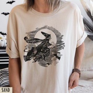 May include: Sand-colored t-shirt featuring a black and white illustration of a witch flying on a broomstick in front of a full moon, with a black cat and bats. The word "SAND" is printed at the bottom.
