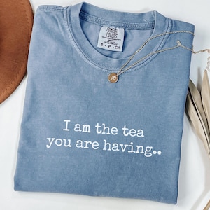 May include: A blue Comfort Colors t-shirt with the text "I am the tea you are having." A gold necklace with a pendant rests on the shirt. The shirt is folded and has a tag at the neckline.
